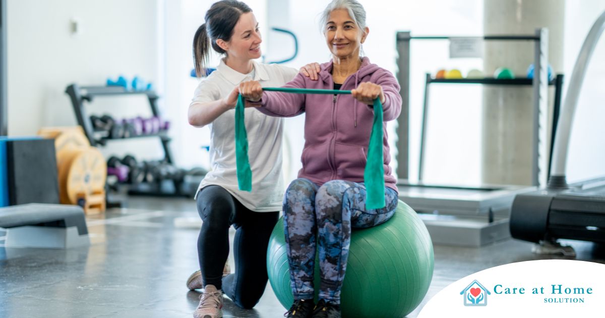 A care provider helps an older woman exercise with a resistance band and an exercise ball, representing how exercise can help with senior fall prevention.
