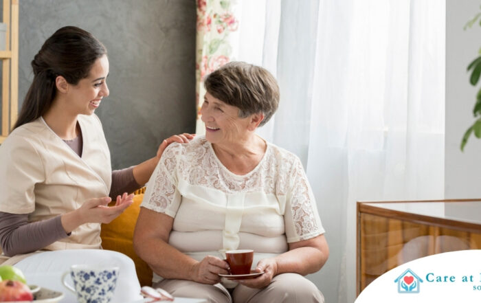 Smiling caregiver with her hand on an older woman’s shoulder as they share tea on the couch, showing home care vs assisted living