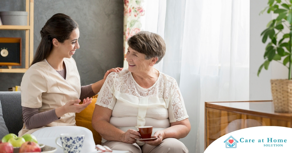 Home Care vs Assisted Living Where Caregiving Work Fits in Today’s Families Smiling caregiver with her hand on an older woman’s shoulder as they share tea on the couch, showing home care vs assisted living